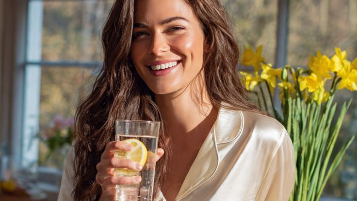 Woman in satin pajamas holding a glass of water with lemon slices in a kitchen.